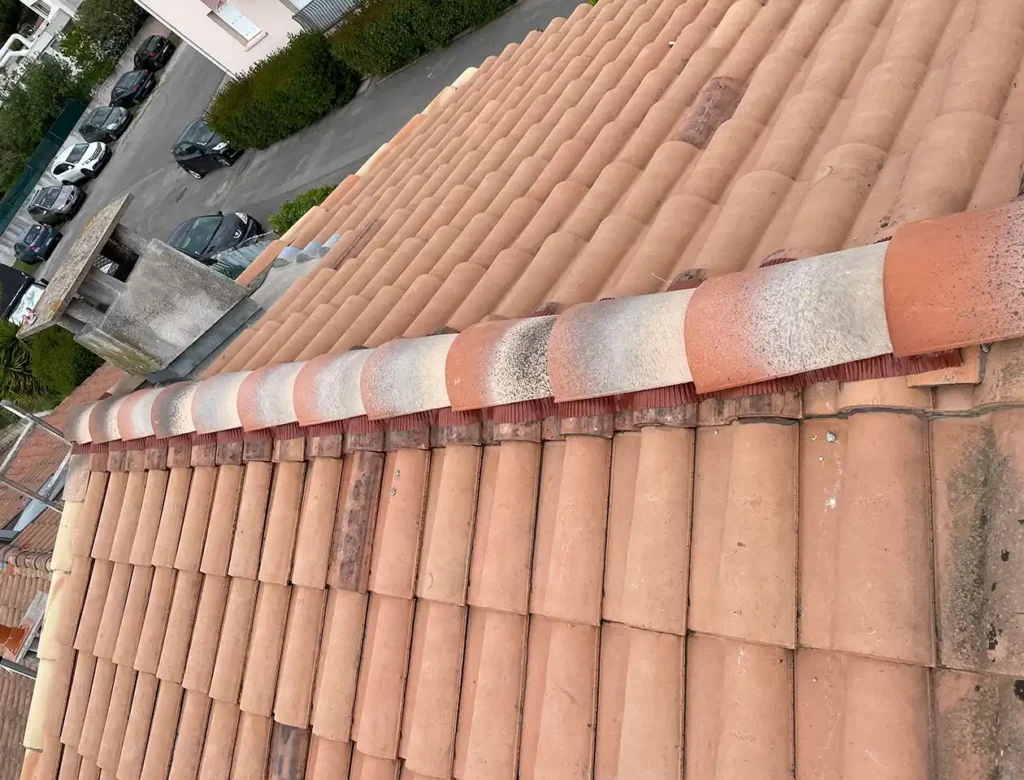 Une vue rapprochée d'un toit en tuiles de terre cuite met en évidence l'alternance des couleurs sur les tuiles faîtières après un changement de faitage, avec une cheminée et des voitures en stationnement visibles à l'arrière-plan.