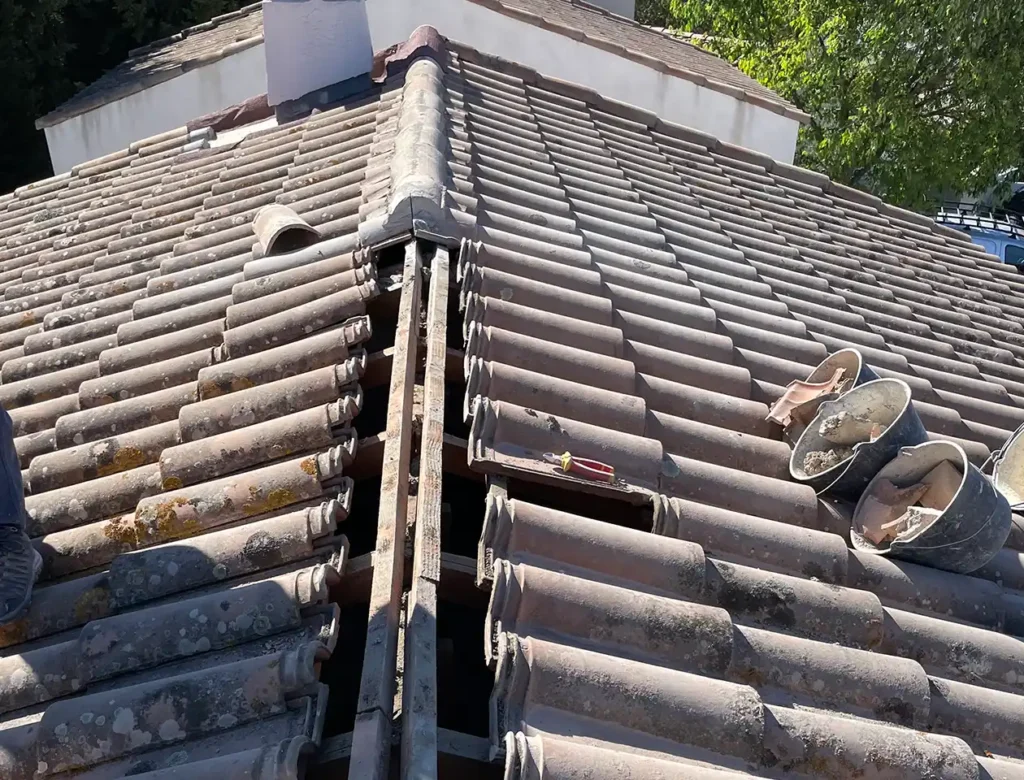 Un toit de tuiles avec une crête centrale visible où des tuiles sont manquantes pour réparation, ce qui indique un changement de faîtage. Des outils et des chaussures sont placés sur le toit, avec une partie d'un bâtiment blanc et des arbres verts en arrière-plan.