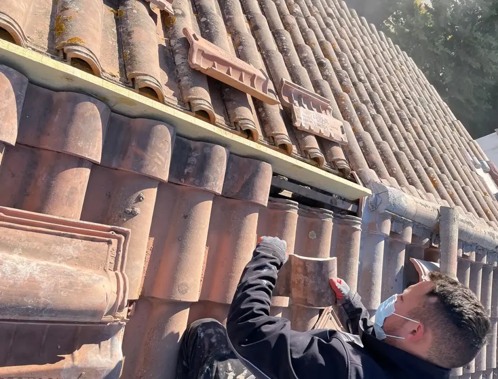 Une personne portant un masque répare un toit de tuiles, travaillant sur un changement de faîtage, tenant une tuile et un outil, avec plusieurs tuiles enlevées et empilées à proximité. La lumière du soleil projette des ombres sur les tuiles.