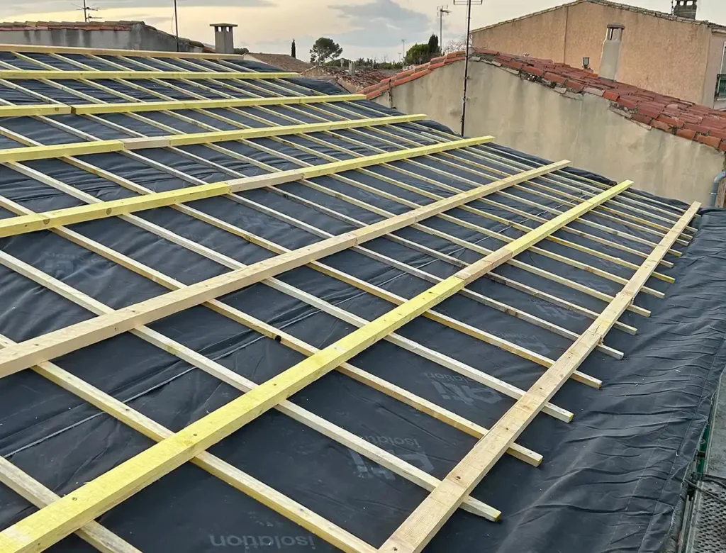 Toit en construction illustrant la création de toiture neuve, avec des lattes de bois disposées en grille sur un matériau d'étanchéité noir, prêtes à recevoir les tuiles, et les toits voisins visibles à l'arrière-plan.