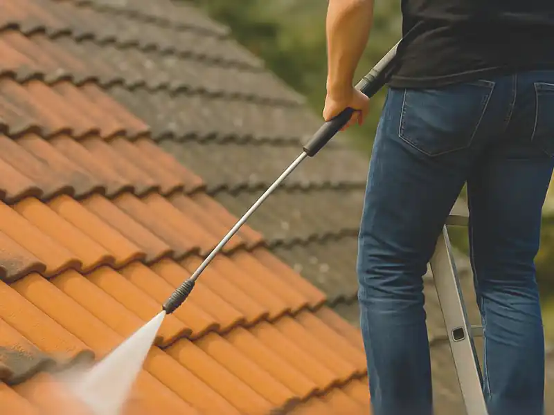 Nettoyage basse pression Une personne debout sur une échelle utilise un nettoyeur à pression pour un nettoyage basse pression, en pulvérisant de l'eau sur les tuiles orange pour les nettoyer en douceur.