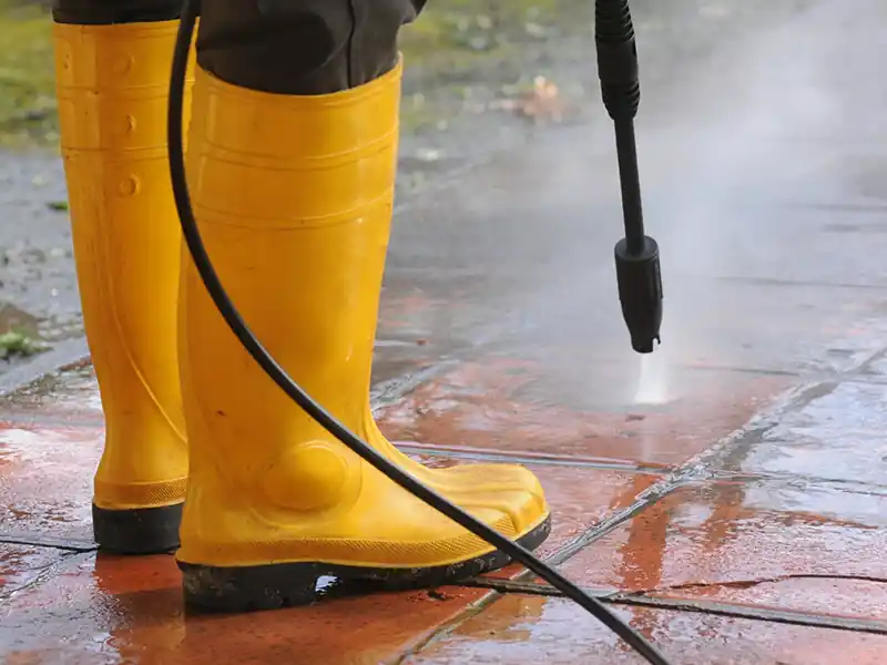 Nettoyage cours et allées - Renovation Sud Personne portant des bottes en caoutchouc jaunes utilisant un nettoyeur à pression pour nettoyer des tuiles rouges à l'extérieur, avec un jet d'eau visible sur le sol-idéal pour le nettoyage cours et allées.