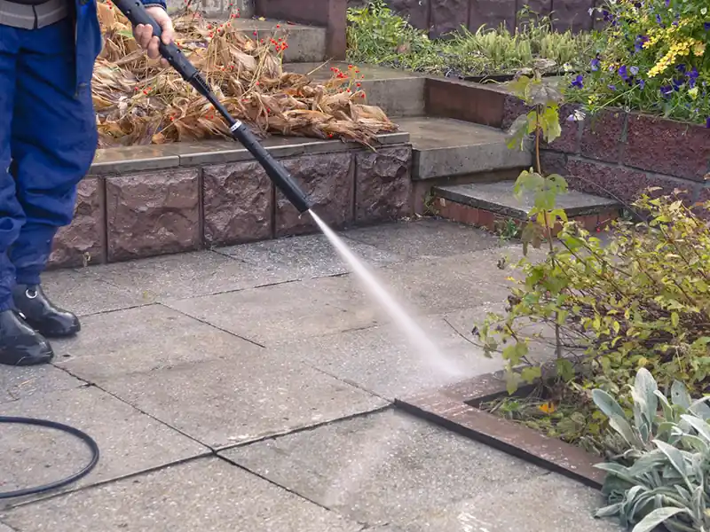 Nettoyage dalles béton - Renovation Sud Une personne utilise un nettoyeur à pression pour le nettoyage extérieur des dalles béton sur une terrasse ou une allée en pierre dans un jardin, avec des plantes et des marches visibles à l'arrière-plan.