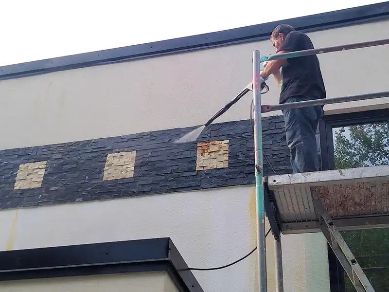 Nettoyage de hangar et entrepot - Renovation Sud Une personne montée sur un échafaudage utilise un nettoyeur à pression pour nettoyer les carreaux de pierre du mur extérieur d'un bâtiment moderne, selon des techniques similaires à celles utilisées pour le nettoyage de hangar et d'entrepôt.