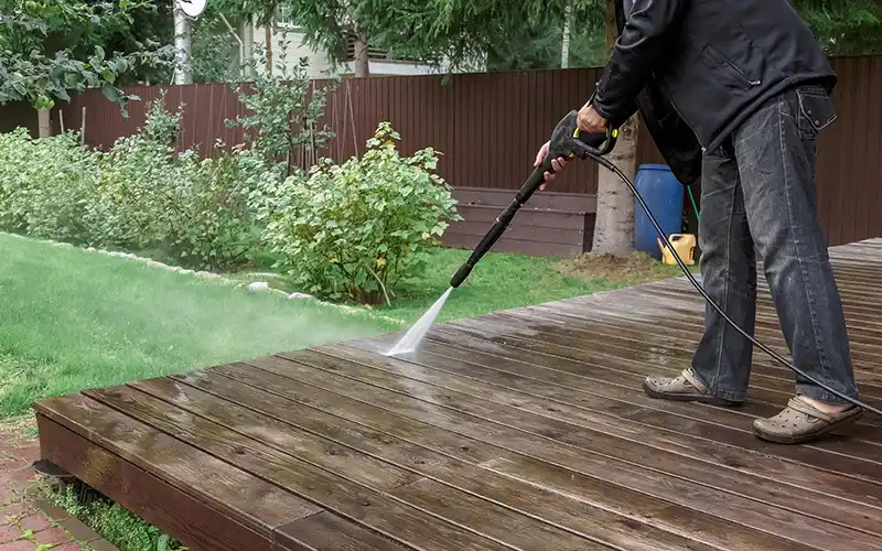 Nettoyage extérieur - terrasses, allées, cours - Renovation Sud près de Nimes Une personne vêtue d'une veste noire et de chaussures grises utilise un nettoyeur haute pression pour nettoyer l'extérieur d'une terrasse en bois dans une arrière-cour avec de l'herbe verte, des arbustes et une clôture marron.
