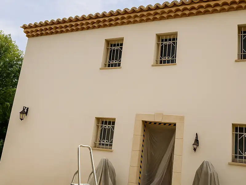 Peinture de facade Nimes - Renovation Sud Un bâtiment en stuc beige, probablement en cours de peinture de façade, possède quatre petites fenêtres grillagées et une porte recouverte d'une bâche en plastique. Deux objets recouverts de tissu sont posés près de l'entrée, et une échelle métallique se trouve au premier plan.