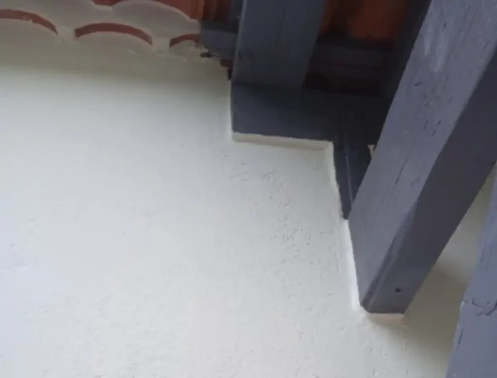 Vue rapprochée d'un mur en stuc blanc avec peinture sur façade, rencontrant un plafond avec des poutres apparentes en bois foncé et quelques tuiles courbes visibles dans le coin supérieur gauche.