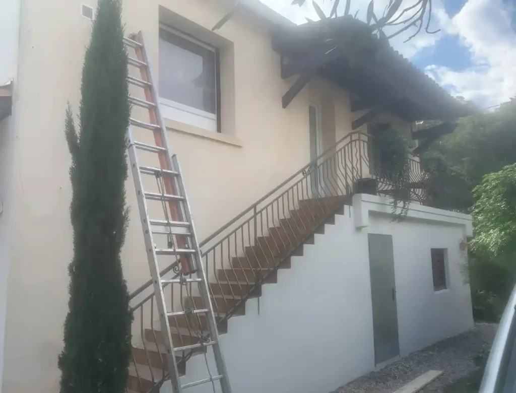 Une échelle métallique s'appuie sur le mur extérieur d'une maison à deux étages, à côté d'un escalier extérieur, suggérant une peinture de façade récente. Des arbres et de la verdure entourent la maison sous un ciel partiellement nuageux.