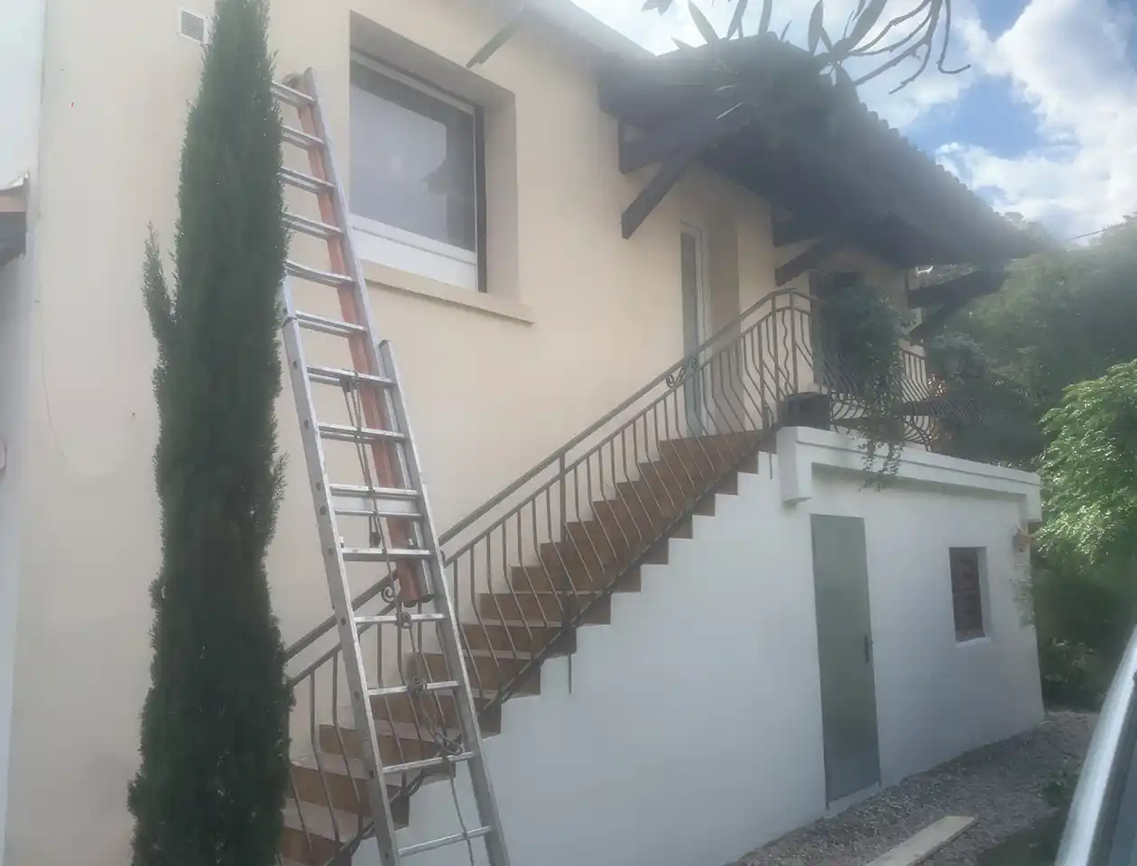 Une échelle métallique s'appuie sur le mur extérieur d'une maison à deux étages, à côté d'un escalier extérieur, suggérant une peinture de façade récente. Des arbres et de la verdure entourent la maison sous un ciel partiellement nuageux.