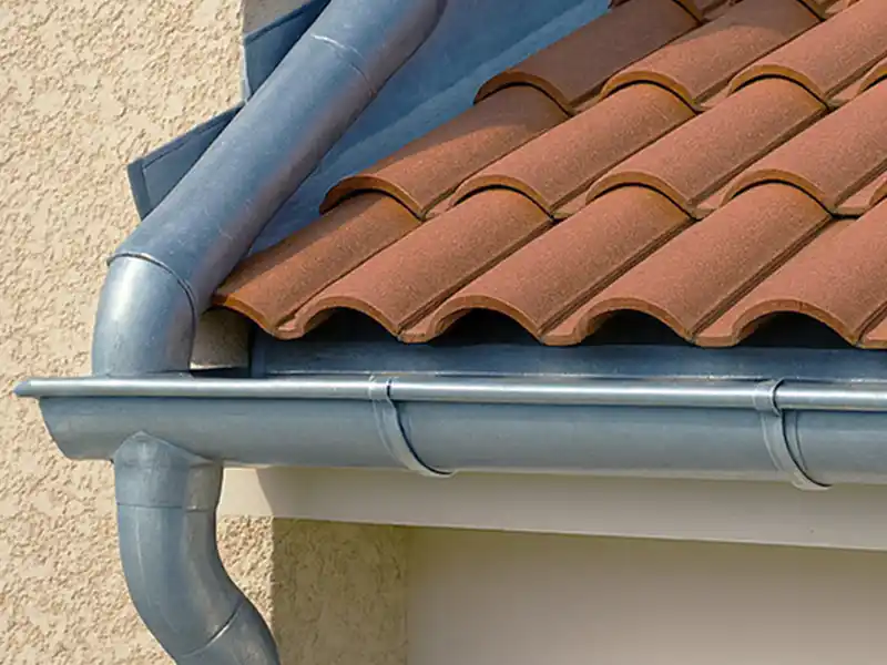 Pose et nettoyage de gouttière dans le Gard - Rénovation Sud Gros plan sur le toit d'une maison aux tuiles rouges courbes en terre cuite et au système de gouttière en zinc, montrant la descente d'eau fixée au mur extérieur beige - un exemple de pose de gouttière dans le Gard.