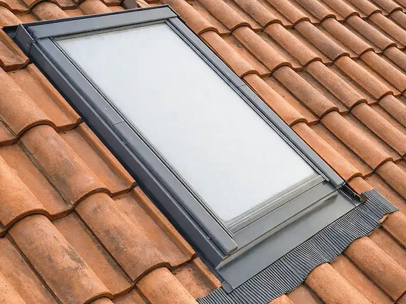 Pose fenêtre de toit à Nimes et dans le Gard - Rénovation Sud Une lucarne rectangulaire avec un cadre noir est installée sur un toit avec des tuiles en terre cuite orange. La fenêtre de toit semble fermée et est entourée d'un solin de protection, idéal pour la pose fenêtre de toit à Nîmes et dans le Gard.
