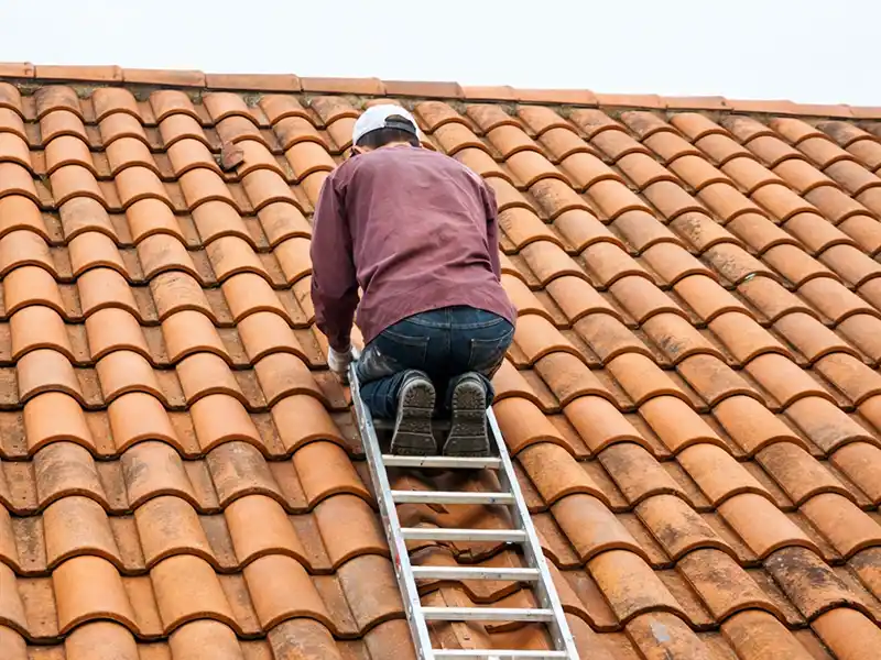 Réparation de toiture dans le Gard 30 - Rénovation Sud Une personne s'agenouille sur un toit incliné en tuiles orange, monte sur une échelle, porte une casquette blanche, une chemise à manches longues marron et un jean, et semble inspecter ou effectuer une réparation de toiture dans le Gard.