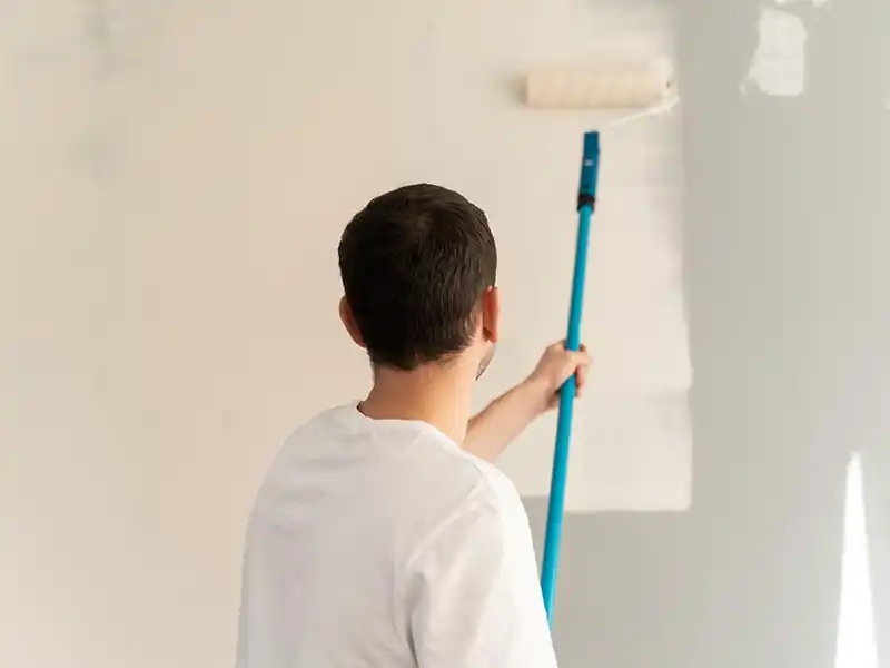 Travaux de peinture intérieure - Rénovation Sud à Nimes Une personne portant une chemise blanche effectue des travaux de peinture intérieure, en utilisant un rouleau de peinture avec un manche bleu pour transformer un mur de gris en blanc. Le mur est partiellement peint et la personne est vue de dos.
