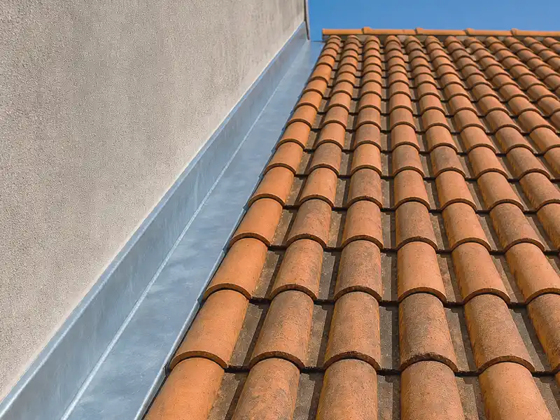 Travaux de zinguerie et étancheité dans le Gard - Rénovation Sud Vue rapprochée d'un toit de tuiles en terre cuite à côté d'un mur en stuc beige, avec des travaux de zinguerie et un solin métallique le long du bord où le toit rencontre le mur, sous un ciel bleu clair.